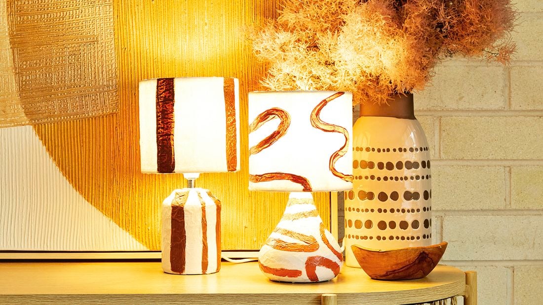 Two abstract paper mache lamps with lights on next to a vase on a table in front of an artwork and brick wall