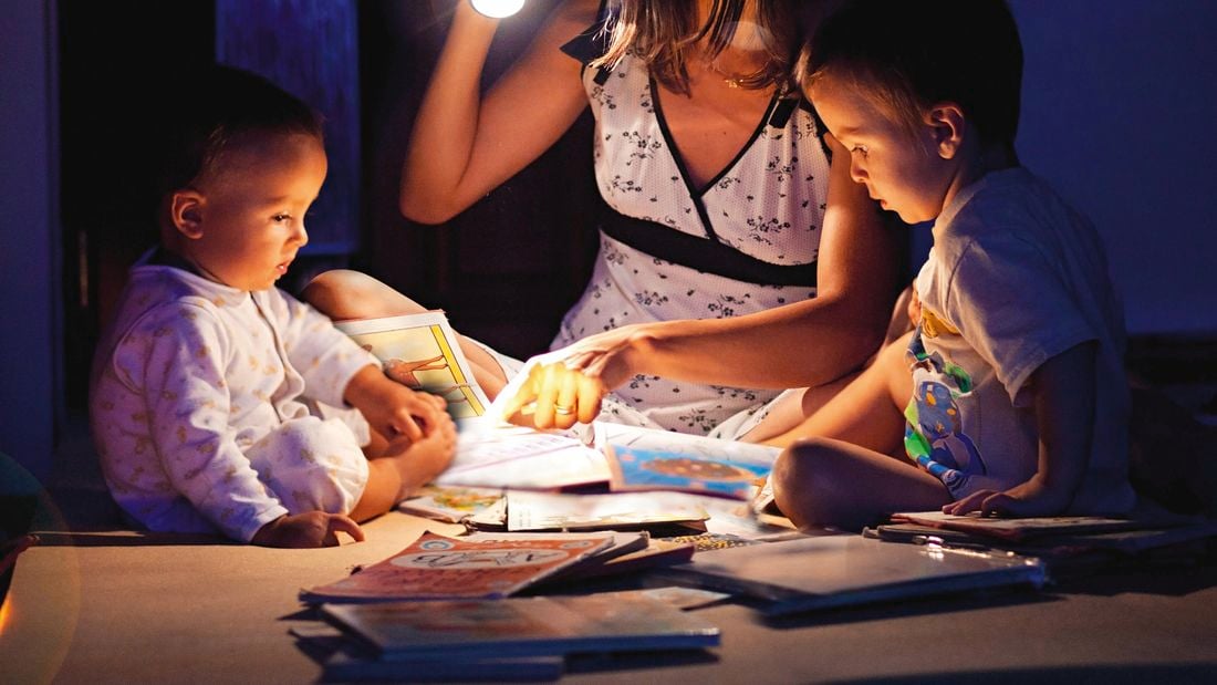 A mother and her two children read storybooks by torchlight on the floor.