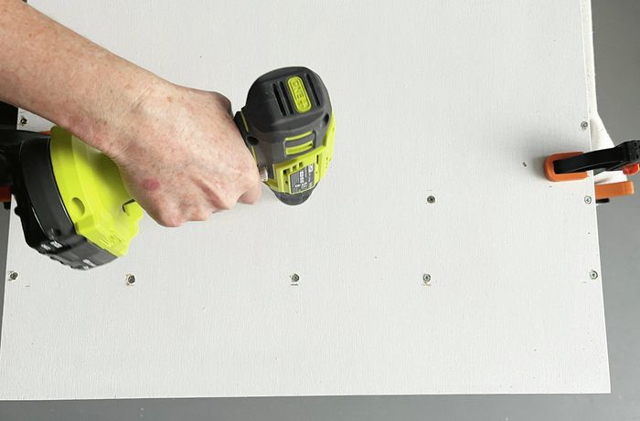 A hand using a drill to make screw holes on a clamped white-painted plywood panel. [[2609] MAK...me sign_V1 | Word]