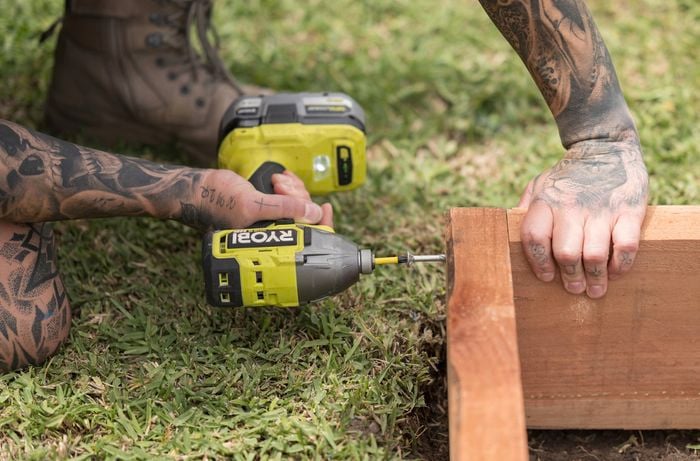 A person is using a Ryobi power drill to fasten a screw into a sleeper to form the frame while keeping it steady on the grass.