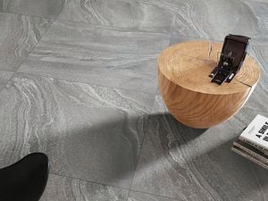 Grey tiled flooring, a wooden round coffee table with a camera on top, and a stack of books on the floor. Part of a black chair is visible in the foreground.