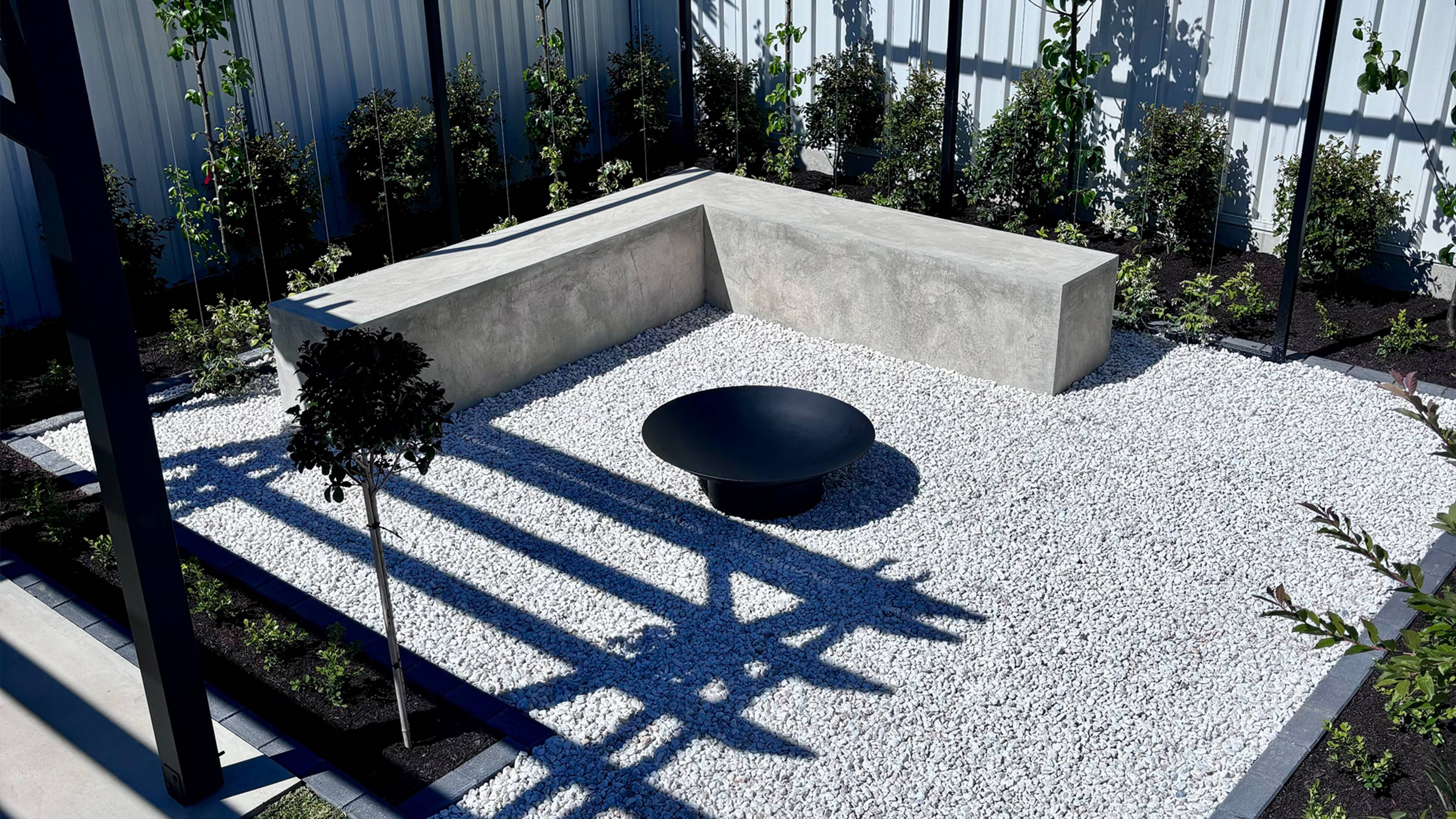 A landscaped yard with a black fire pit sitting on pebbles, with a cement corner bench seat and hedging