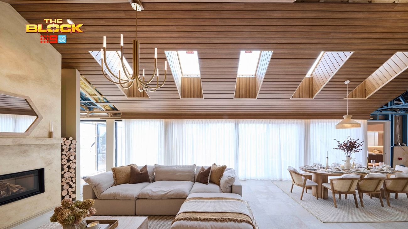 A modern, open-plan living and dining area with a wooden ceiling and multiple skylights. The living space features a large beige sectional sofa with cushions, a wooden coffee table, and a fireplace on the left. The dining area includes a long table set for eight with a pendant light overhead. Floor-to-ceiling curtains line the back wall, letting in natural light. 