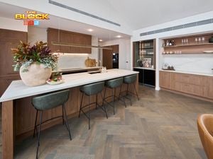 A modern white kitchen with brown cupboards, bar stools at the benchtop with a vase with flowers in it and two bowls with items in them on it.