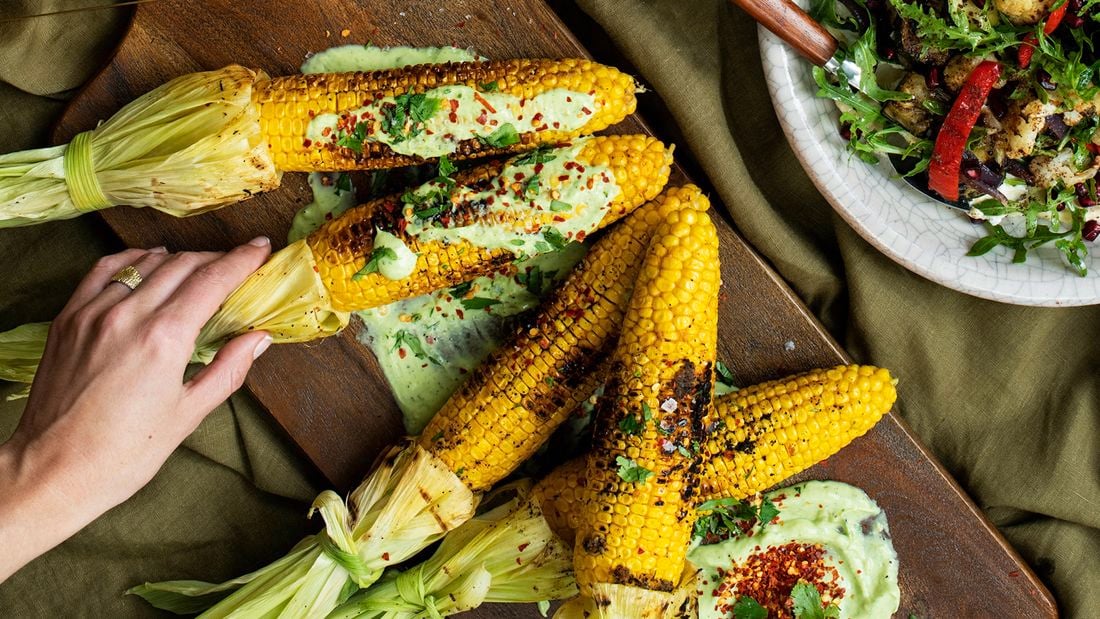 A wooden serving board with BBQ corn on the cob sitting on avocado sauce and topped with lime butter, chill and coriander