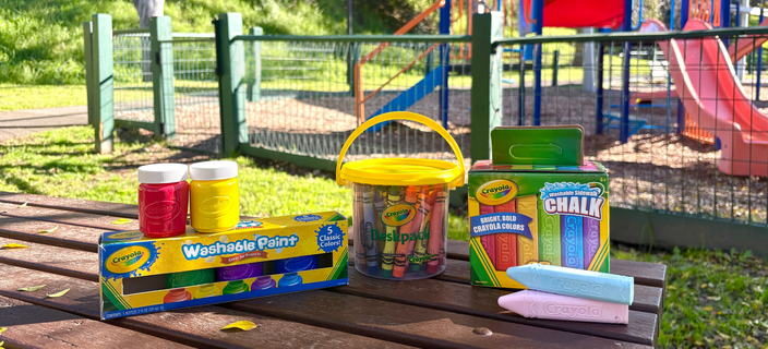 Selection of Crayola art and craft products, including washable paint, crayons and sidewalk chalk, displayed on an outdoor picnic table near a playground.