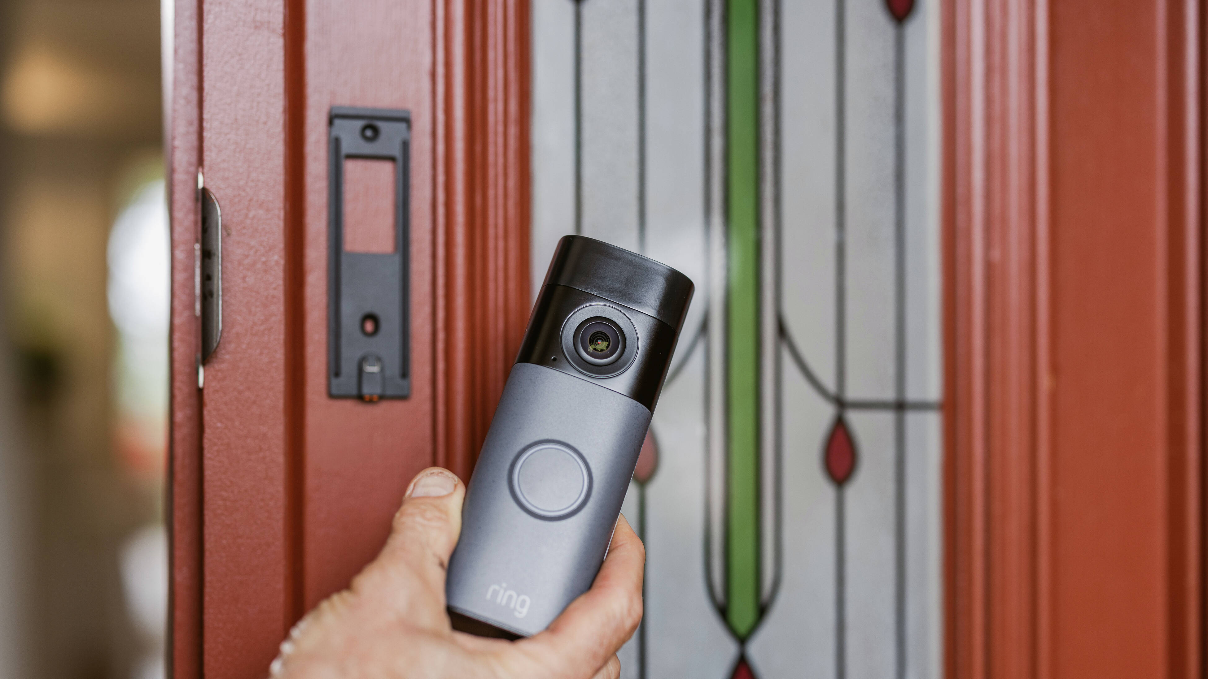 Hand holding a Ring video doorbell camera being installed on a red front door with decorative glass panel.”
