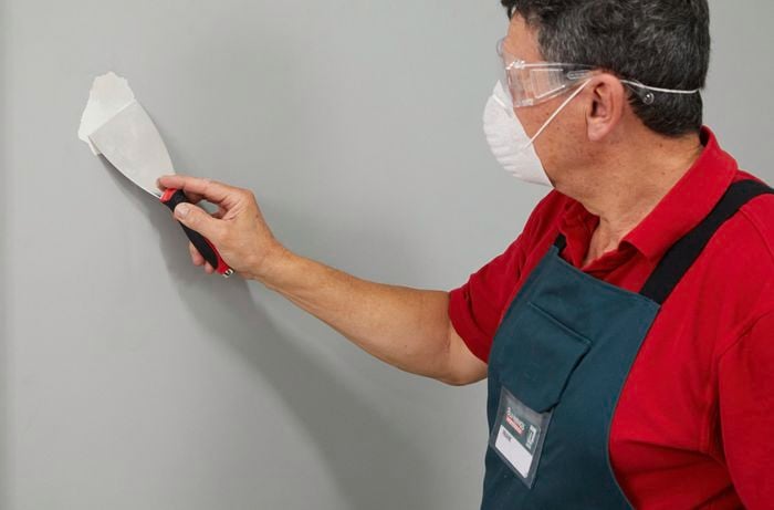 A man wearing a dust mask and safety glasses smooths a wall with a paint scraper.