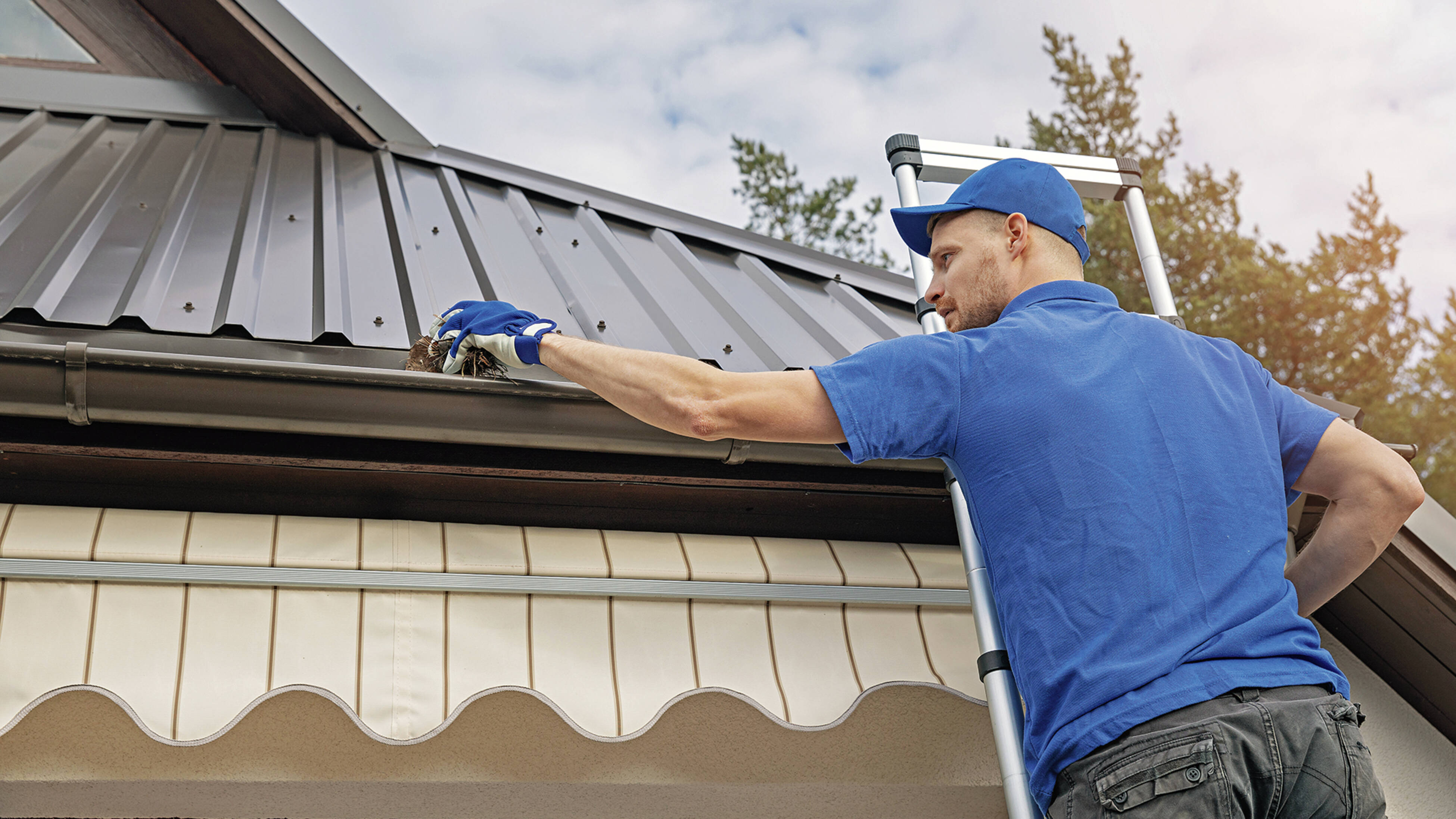 A man wearing a blue shirt and gloves stands on a ladder, cleaning debris from a house gutter under a metal roof to guard against extreme weather.