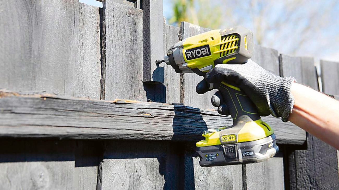 A hand holding a Ryobi cordless drill, drilling a hole into a grey outdoor wooden fence