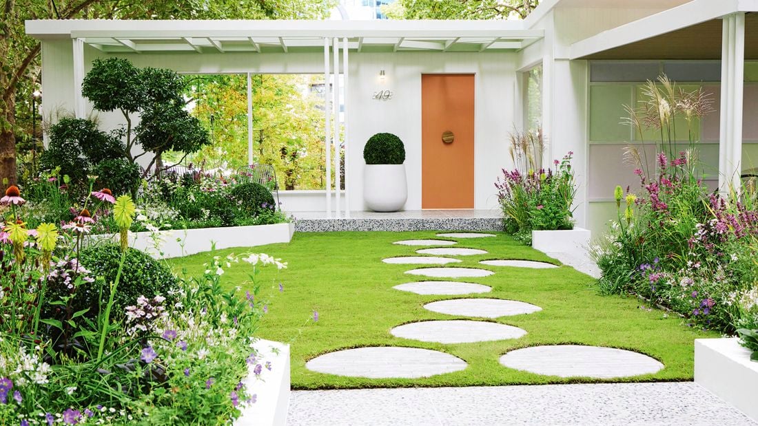 A well-manicured garden with green grass, white stepping stones, white edging, green trees and blooming flowers