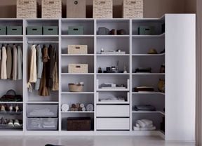 A large open wardrobe unit with shelves and hanging rails holding clothes, shoes, folded towels, storage boxes, wicker baskets, drawers and boxes on top. A floor lamp stands on the left.