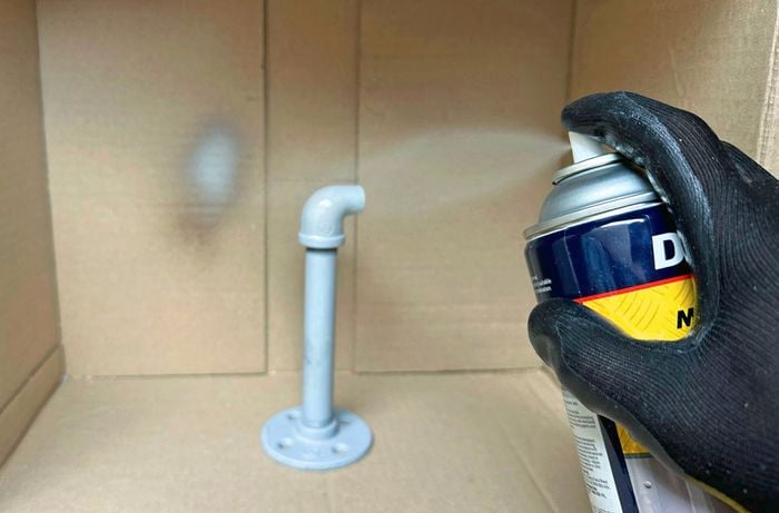 A black gloved hand spray painting a pipe with metal primer using a cardboard box as a spray booth