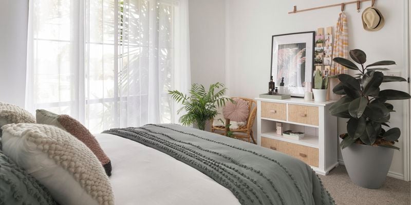 Bedroom with sheer curtains, bed, chest of drawers and potted plants.