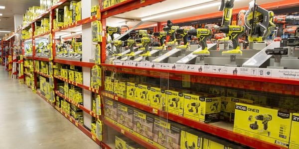 Hardware store aisle displaying Ryobi power tools and accessories on red shelving. Multiple cordless drills and saws are mounted on display racks, with rows of bright yellow product boxes stocked on shelves below and above.