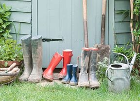 Gardening tools and equipment next to a shed, including gumboots, a shovel, a pitchfork and a watering can.