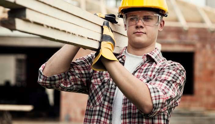 A tradesman wearing a yellow hard hat carrying several lengths of timber on a construction site