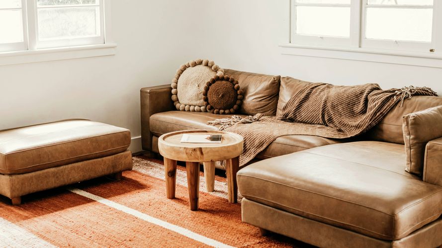 Cozy living room featuring a tan leather sectional sofa, matching ottoman, round timber coffee table and textured rug, illuminated by natural light from large windows.