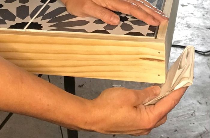 A person uses abrasive paper to sand down the corners of a tiled outdoor ta