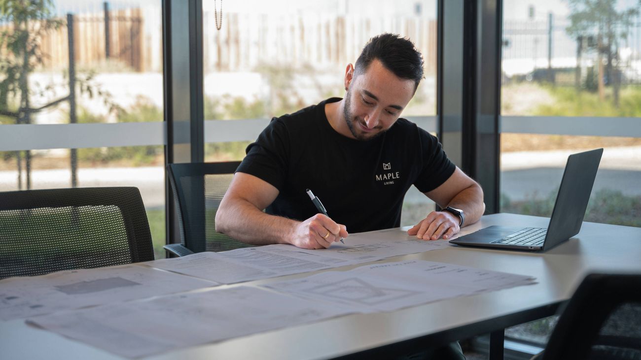 A man sitting at a desk indoors looking down at plans with a pen in his hand and an open laptop in front of him