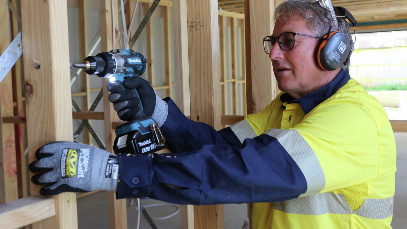 A tradesperson wearing earplugs, safety glasses and Mechanix Wear gloves uses a Makita hammer drill at a partially framed construction site.
