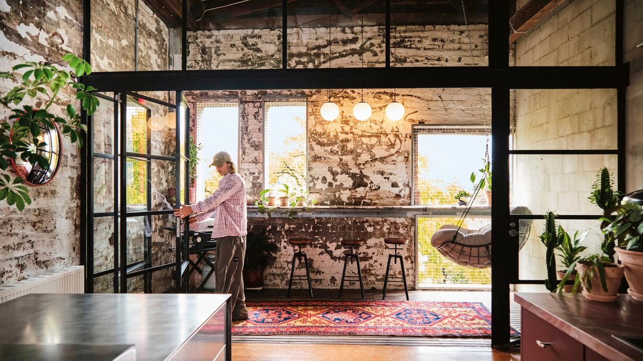 A person in a cap and plaid shirt stands in an industrial-style room with exposed brick, large windows, plants, and a patterned rug
