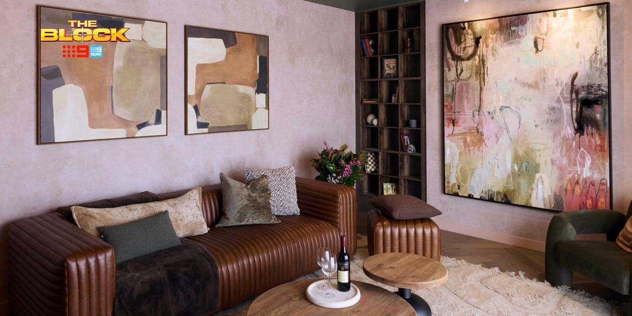 A modern living room featuring a brown leather sofa with assorted cushions, two round wooden coffee tables, and an abstract painting on the wall. Two smaller abstract artworks hang above the sofa. A bookshelf is in the corner, and a green armchair sits to the right. 