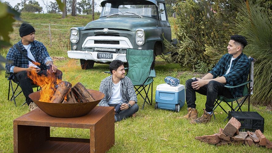 Three friends in front of a fire, one too cool to sit in his camping chair