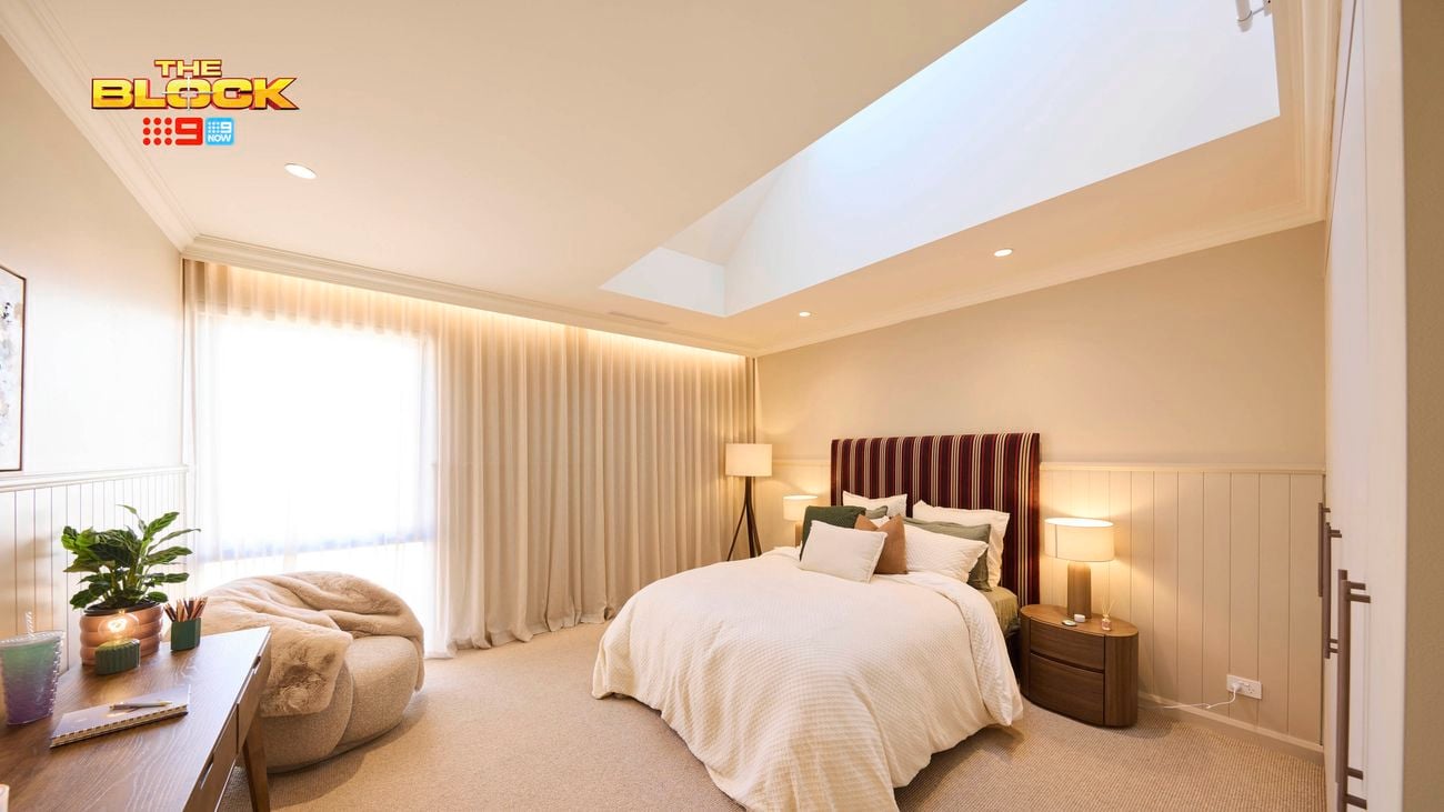Cream bedroom with a double bed and a patterned bedhead, white linens and a lamp in the corner and a bean bag and table on the opposite wall. 