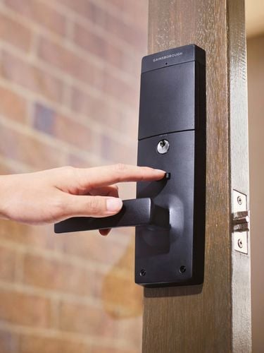 Hand pressing the handle of a black GAINSBOROUGH electronic door lock on a wooden door near a brick wall.