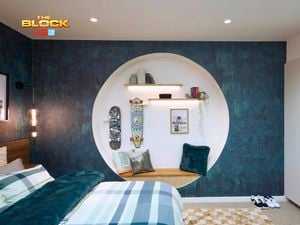 Blue bedroom with a double bed, textured blue wallpaper and blue checked linen. The bed is on a brown checked rug and there is a circular recess in the wall with a bench seat and clothing hooks.