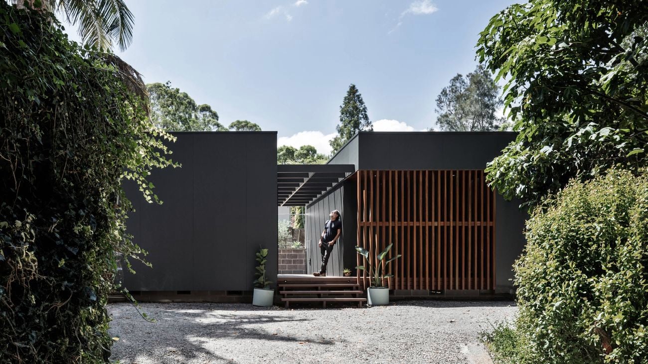 Modern single-storey home with dark cladding and vertical timber batten screening, surrounded by lush greenery, featuring a central entry platform where a person stands beneath a pergola structure.
