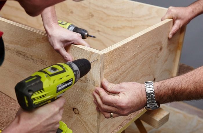 A person drilling a hole through two sheets of plywood joined at right angles