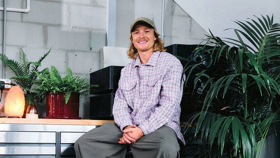 Person sitting indoors beside potted plants and a salt lamp, wearing a cap and patterned jacket.