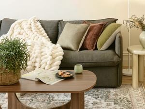 A cosy and warmly lit living room with a sofa with cushions and a throw, a wooden coffee table, shelving and a patterned rug