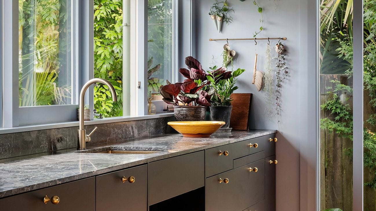 Kitchen with dark grey cabinets, brass knobs, and a marble benchtop; stainless-steel tap beneath three windows looking onto dense greenery. Hanging plants and utensils on the wall, a wooden bowl and potted plant on the bench, light timber floor, and a tall glass door to the garden.