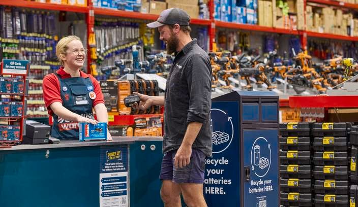 Customer at a Bunnings trade counter handing over an 18V battery, with battery recycling signage and power tools displayed in the store.