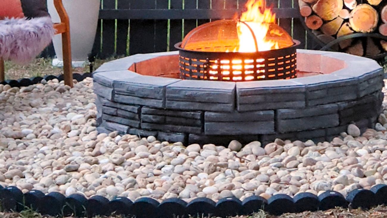 Stone fire pit with burning fire, surrounded by pebbles, stacked firewood and outdoor seating.