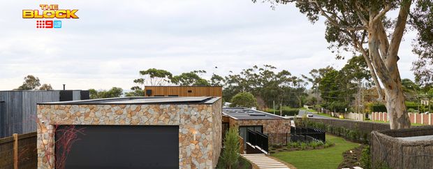 Modern garage with natural stone cladding, a black roller door, curved stepping stone pathway, landscaped lawn and mature trees.