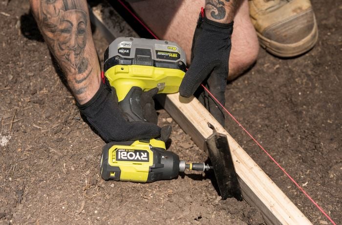 A Bunnings Team Member is using a Ryobi power drill to secure a temporary border, aligning it with a red string line to keep your base intact while you work. 