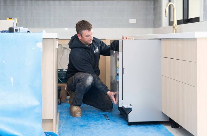 A tradesperson is installing a dishwasher in an existing kitchen layout with light wood cabinetry and a brass tap.