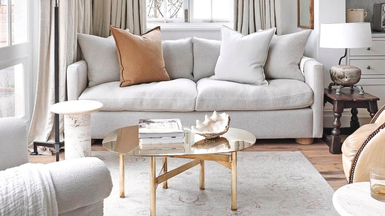 Grey couch with grey curtains and flooring in a living room. Other furnishings like a coffee table, a white lamp and a large window are also featured