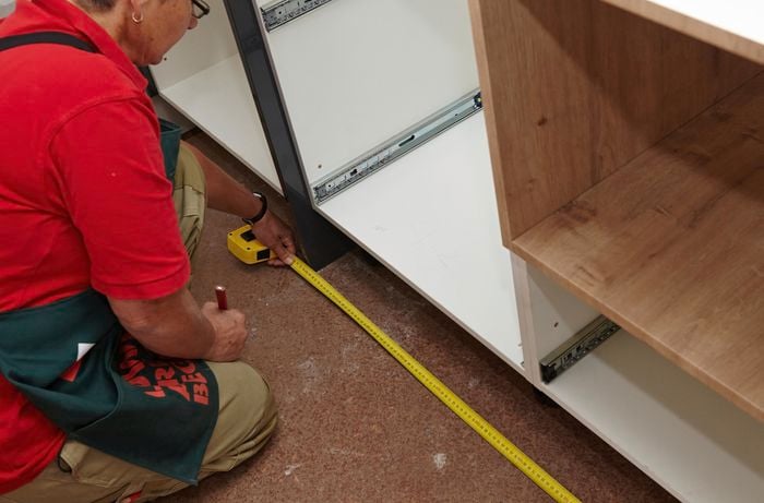 A person kneeling on the floor measuring the length of a cabinet