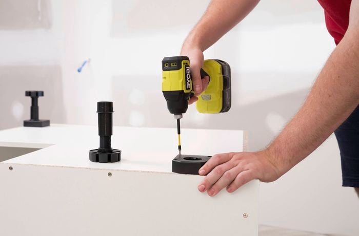 A person uses a power drill to attach feet to the bottom of a corner cabinet.