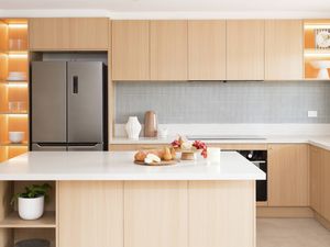 This image shows a clean kitchen with light wood cabinets, a big island bench, open shelves with warm lighting, and a stainless steel fridge. 