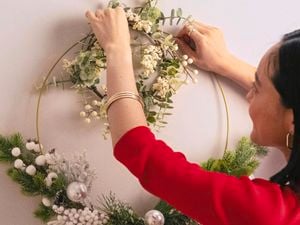 A woman with a dark bob and red top hangs a wreath on the wall using clear Command hooks