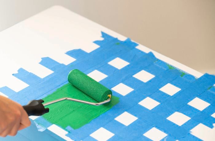 A person is painting the second set of squares on the toy box lid over a taped grid using a paint roller