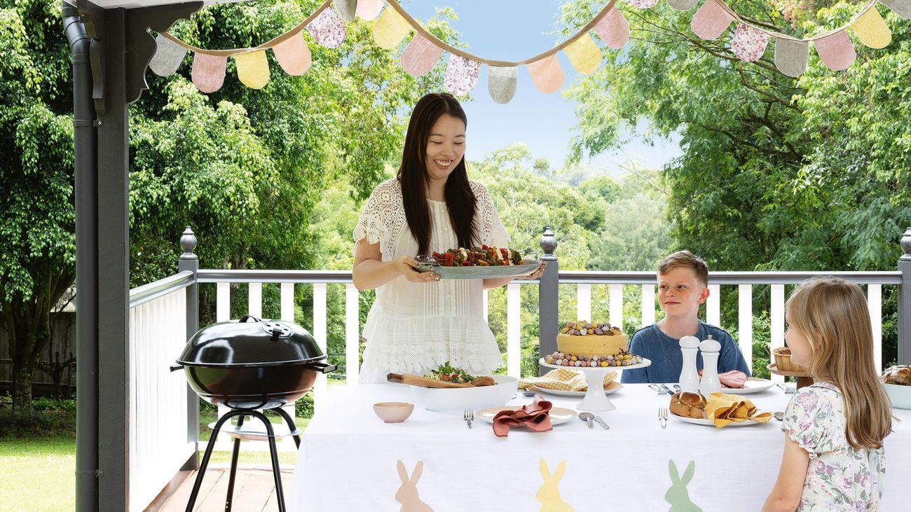 A smiling woman on a verandah in an Easter setting carries a tray of kebabs as two children look on