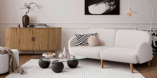 Modern living room featuring a curved white bouclé sofa, glass coffee table with sculptural black sphere bases, light timber sideboard, abstract wall art and an arched floor lamp. The space is styled with neutral tones, textured furnishings and contemporary décor on a herringbone wood floor.