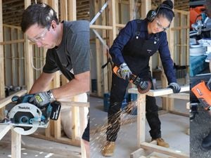 Three workers wearing safety glasses and hearing protection use power tools to cut and shape wood inside a timber-framed building.